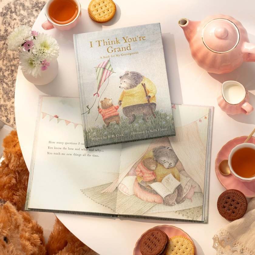 Book titled 'I Think You're Grand' on a table with tea, cookies, and a teddy bear.