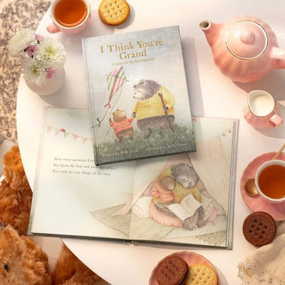 Book titled 'I Think You're Grand' on a table with tea, cookies, and a teddy bear.