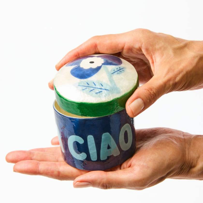 Hand holding a small blue container with a white lid featuring a bird design and 'CIAO' text on a white background.