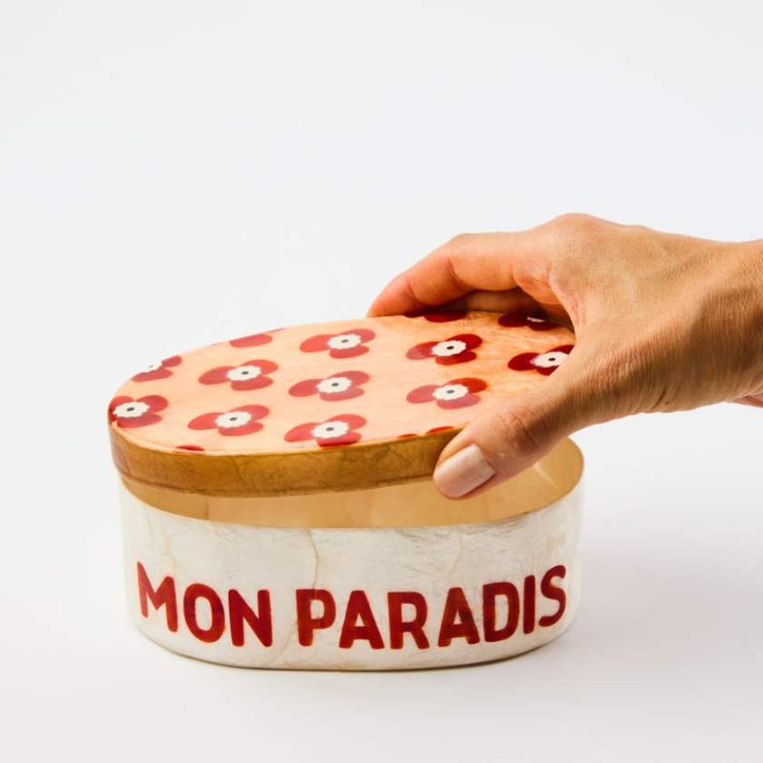 Hand holding a decorative box with floral lid and 'MON PARADIS' text on a white background