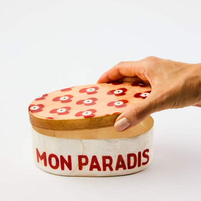 Hand holding a decorative box with floral lid and 'MON PARADIS' text on a white background