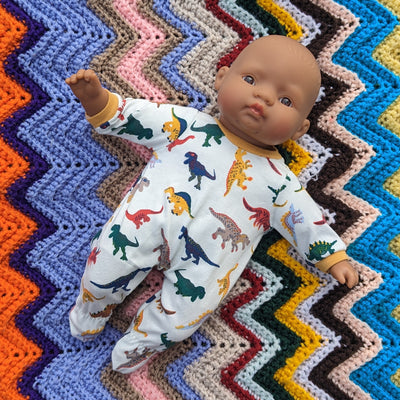 A 32cm soft body hispanic Miniland doll wearing a onesie with white background and colourful dinosaurs