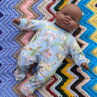 A 32cm soft body African miniland doll wearing a blue onesie with May Gibbs gumnut baby design