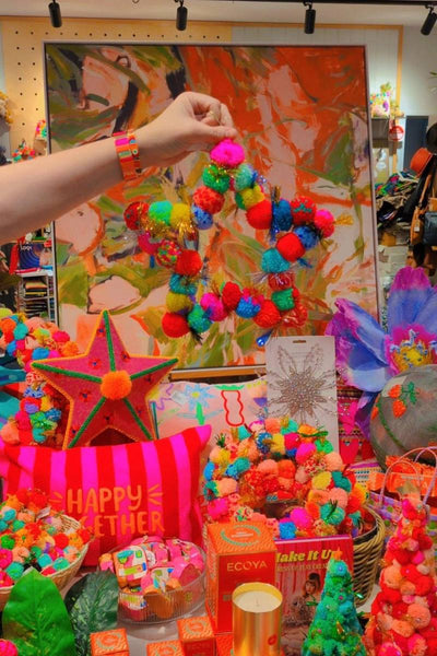Colourful pom-pom wreath being held in front of a vibrant painting with various decorative items | Star pom pom wreath by Arissa available at Zebra Finch Style