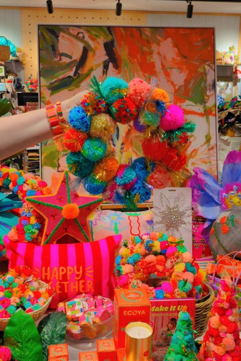 Colorful wreath held by a hand with a vibrant painting in the background | Arissa rainbow pom pom wreaths available at Zebra Finch in Newcastle, NSW