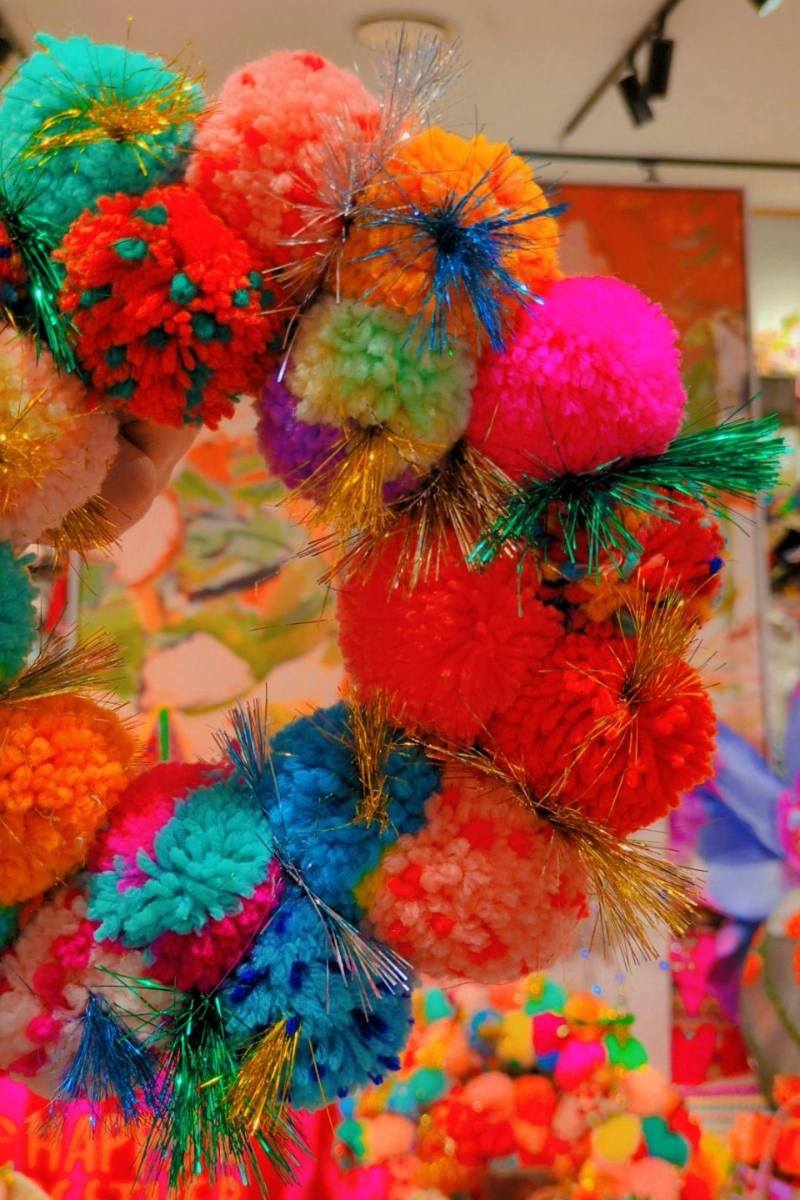 Colorful wreath held by a hand with a vibrant painting in the background | Arissa rainbow pom pom wreaths available at Zebra Finch in Newcastle, NSW