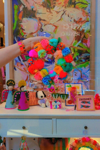 Colorful wreath held by a hand with a vibrant painting in the background | Arissa rainbow pom pom wreaths available at Zebra Finch in Newcastle, NSW