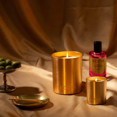 Two gold candles and a pink bottle labeled 'ECOYA' on a beige fabric background.