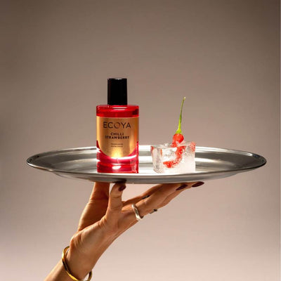 Bottle of ECOYA Chilli Strawberry candle on a metal tray with a small glass of red liquid and a cherry.