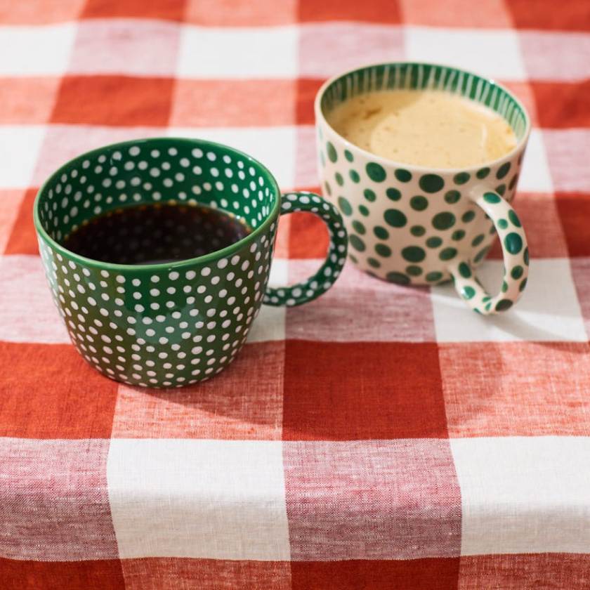 Two green polka dot mugs with one containing a dark liquid and the other a light liquid, on a red and white checkered tablecloth.
