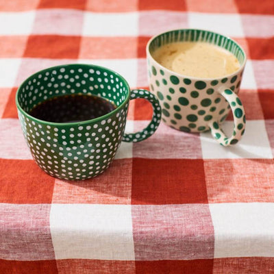 Two green polka dot mugs with one containing a dark liquid and the other a light liquid, on a red and white checkered tablecloth.
