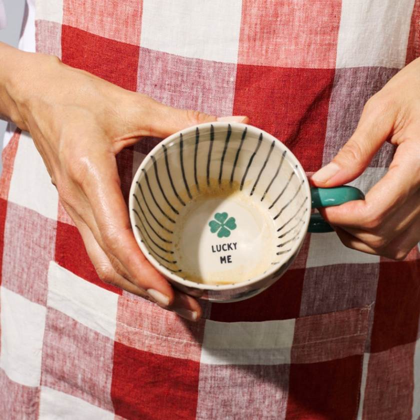 Person holding a ceramic mug with 'Lucky ME' text and clover design against a red and white checkered background.