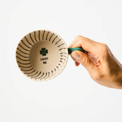 Hand holding a ceramic cup with 'Lucky Me' text and clover design on a white background