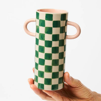 Green and white checkered vase with pink handles held by a hand on a light gray background