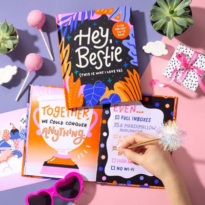 Colorful book titled 'Hey, Bestie' with a hand holding a pen on a pink surface with decorative items.
