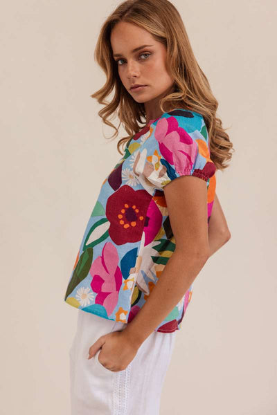 Woman wearing a colorful floral blouse against a beige background. Kaleido linen blouse- stockist at Zebra Finch Marketown in Newcastle, NSW