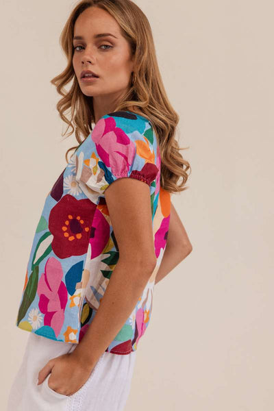 Woman wearing a colorful floral blouse against a beige background. Kaleido linen blouse- stockist at Zebra Finch Marketown in Newcastle, NSW