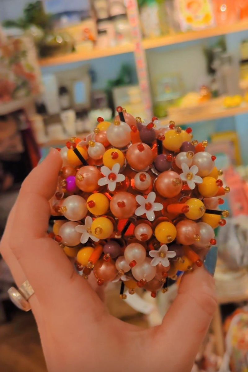 Hand holding a spherical object made of beads and flowers against a blurred indoor background