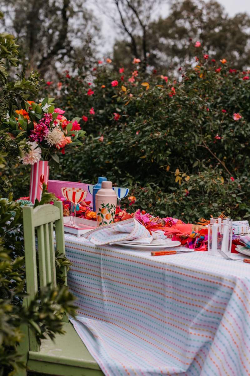 Outdoor table setting with colorful tablecloth, plates, and flowers in a garden.  christmas garland by Mink and Me