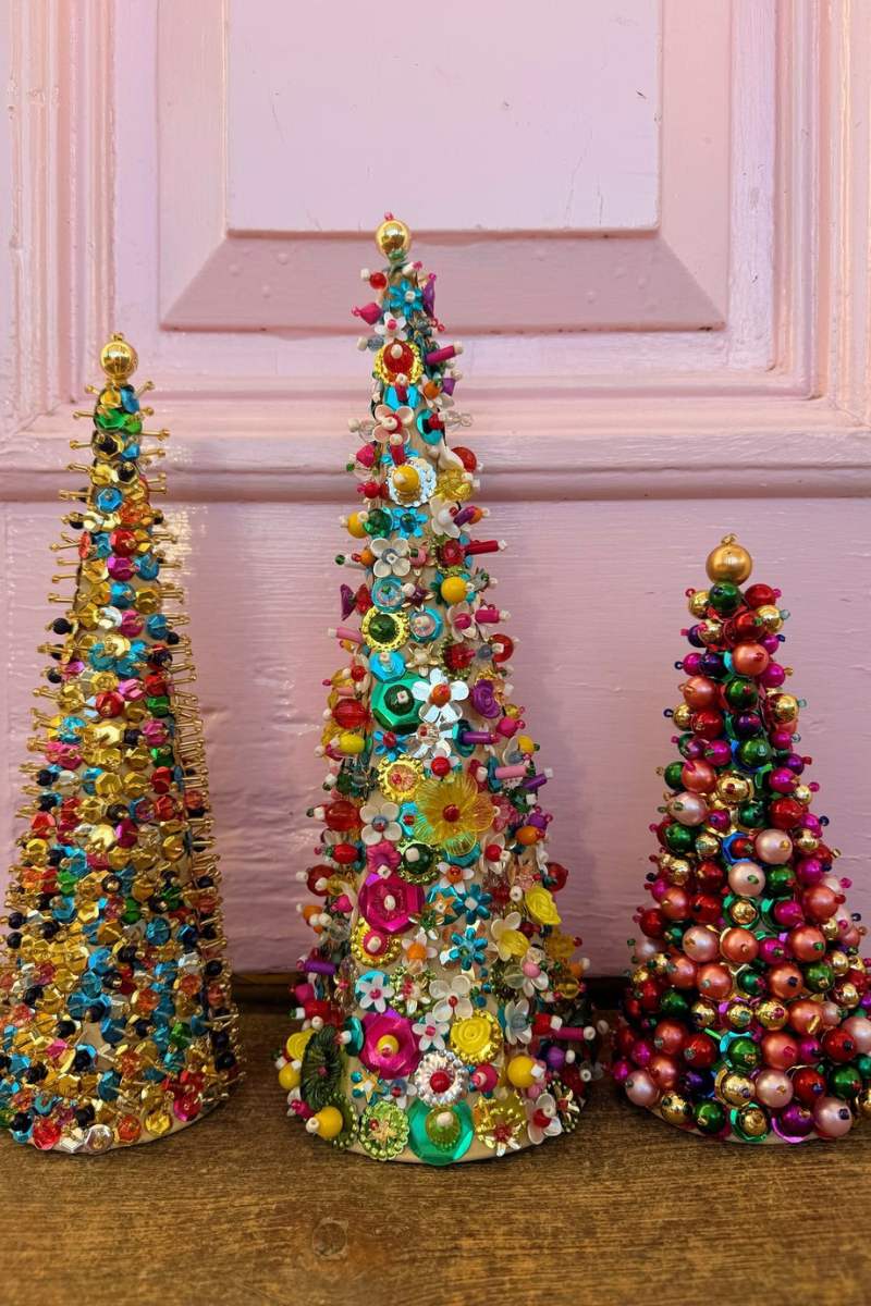 Three colorful beaded Christmas trees on a wooden surface with a pink wall background.