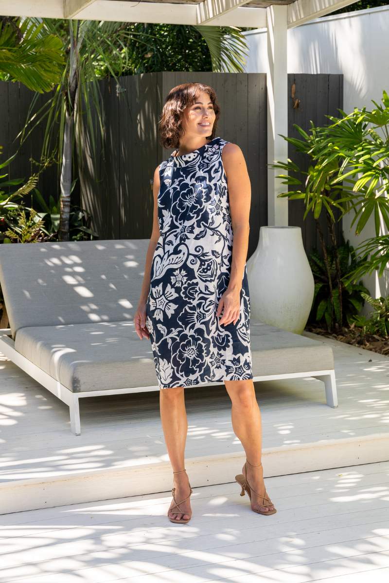 Woman in a navy floral dress standing in a garden setting with plants and a white chair.