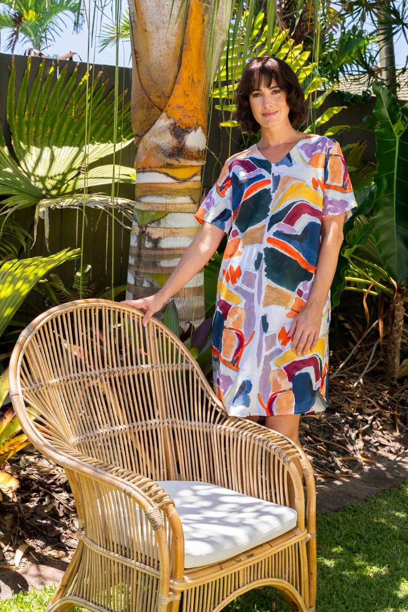 Woman in a colourful dress standing next to a wicker chair with plants in the background