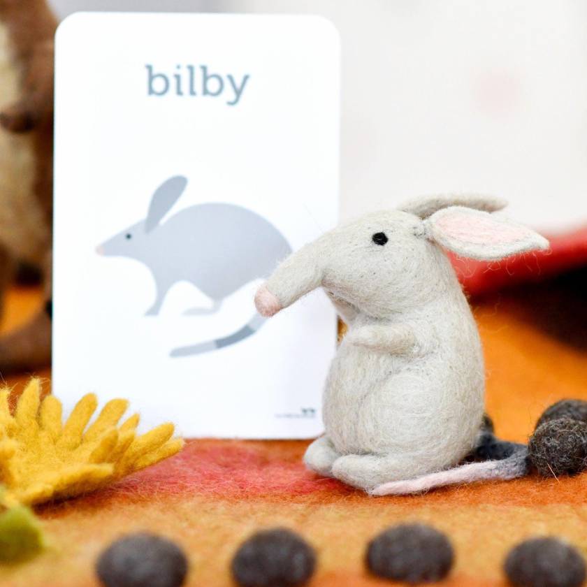 Small plush animal toy resembling a bilby on a colorful surface with a 'bilby' sign in the background.