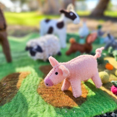 Felted animal toys, including a pink pig, on a green felt surface with a blurred outdoor background.