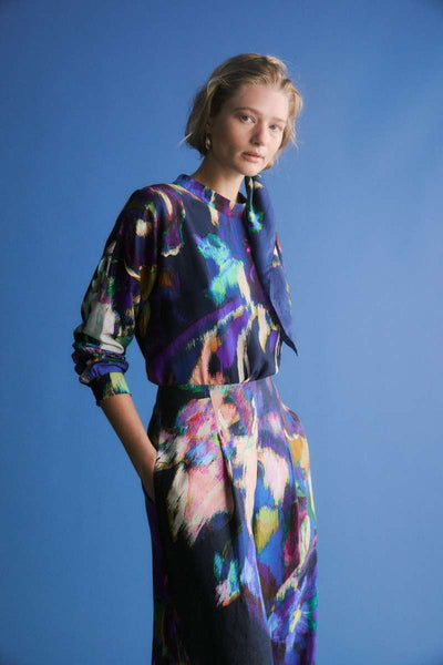 Person wearing a colourful floral outfit against a blue background. Elk the Label Lato Pants in Vaara print, from their 2025 collection. Elk Stockist in Newcastle, NSW at Zebra Finch